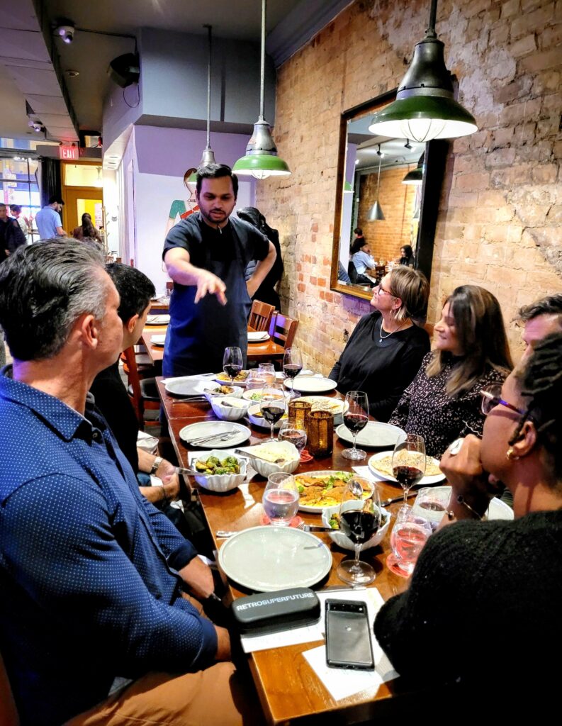 Chef Miheer Shete at Curryish Tavern in Toronto. He is standing at the head of a table of eight people describing the food that they have in front of them.