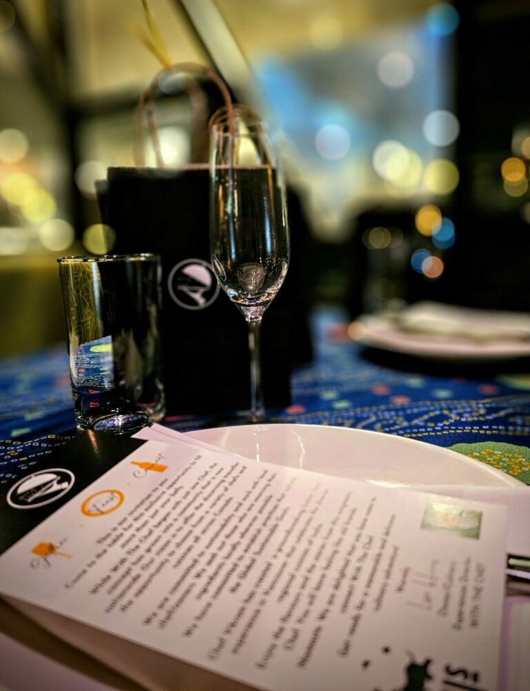 A close up table view with the With The Chef takeaway gift bag, welcome letter and menu.